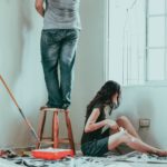 Image of one person standing on a stool and one person sitting on the floor painting the walls of their home during a renovation